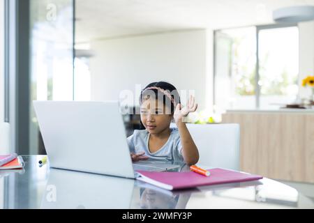 Una ragazza birazziale studia con un laptop in un ambiente luminoso in casa durante una videochiamata Foto Stock