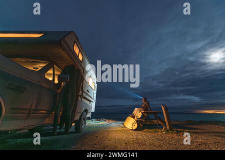 Donna seduta sul tavolo da picnic accanto al camper durante il surftrip Foto Stock