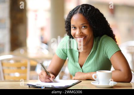 Felice donna nera che compila il modulo in una terrazza del bar che ti guarda Foto Stock