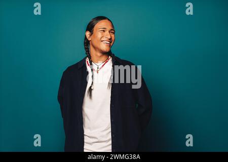 Ritratto di un giovane uomo con capelli intrecciati e gioielli etnici in piedi in uno studio. Uomo che sorride felicemente mentre guarda lontano. I suoi lunghi capelli e la collana Foto Stock
