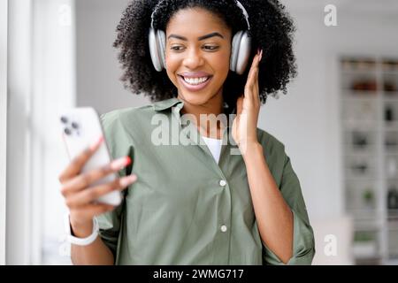 Una vivace donna afro-americana che indossa le cuffie gode di una vivace videochiamata, il suo sorriso illuminato dalla luce del giorno che scorre attraverso una finestra in un ambiente elegante e minimalista da ufficio. Foto Stock