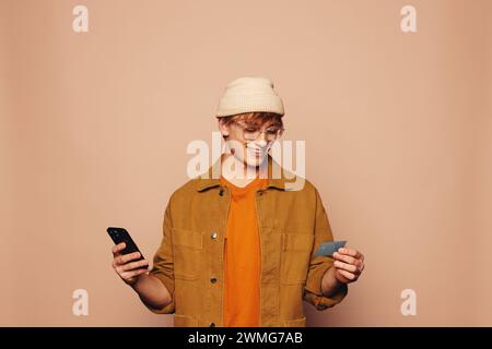 Un giovane uomo felice con giacca casual in denim regge una carta di credito e un cellulare su un vivace sfondo pesca. Rappresenta il mobile banking, lo shopping online Foto Stock