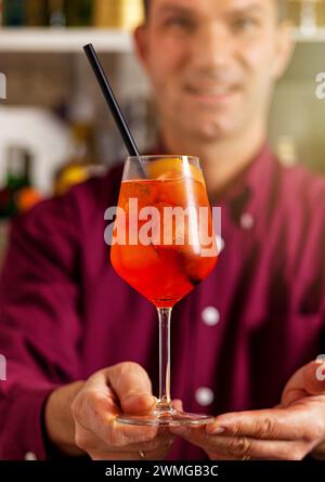 Barman sorridente in una vivace camicia viola che serve un rinfrescante spritz aperol in un bar Foto Stock