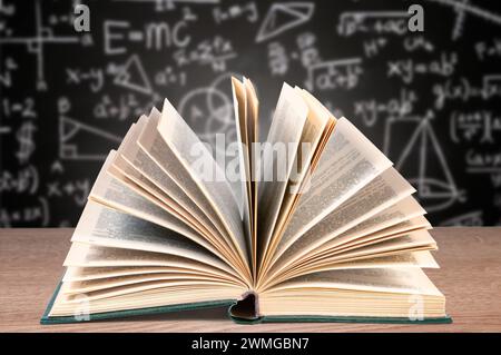Un libro di testo aperto giace su una scrivania di legno in primo piano con uno sfondo sfocato caratterizzato da una lavagna coperta da varie equazioni matematiche Foto Stock