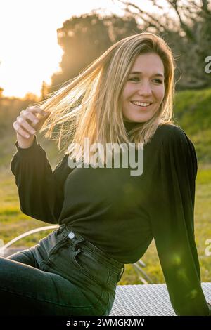 Giovane modella vestita di nero toccando i suoi capelli biondi sorridendo e guardando al suo fianco. Verticale. Foto Stock