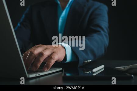 Uomo d'affari in tuta nera che lavora su computer portatile e tablet, digitazione manuale sulla tastiera sul tavolo in ufficio con sfondo scuro, lavoro online, Clos Foto Stock