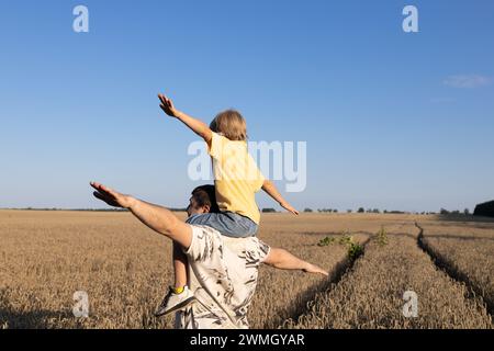 papà e figlio di 6 anni giocano insieme nella natura. l'uomo tiene il bambino sulle spalle, spalmando le braccia come ali di un aereo. Una vacanza in famiglia gioiosa Foto Stock