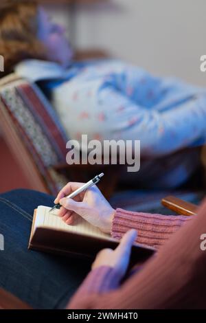 Una psicologa donna adulta intervista una paziente e prende appunti in un quaderno. L'attenzione e' sulle note del medico, e il paziente e' in un BL Foto Stock