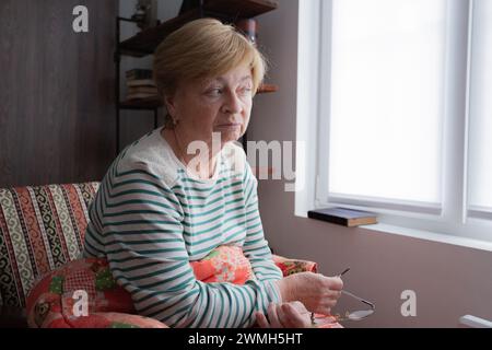 Un'anziana donna caucasica siede su una sedia sotto una coperta vicino alla finestra con gli occhiali in mano e si perde nel pensiero. Foto Stock