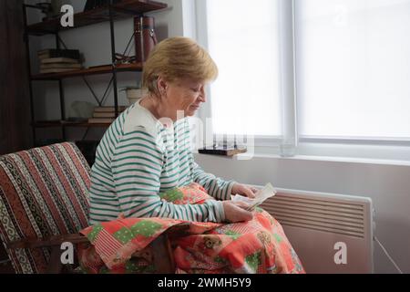 Un'anziana donna caucasica siede su una sedia sotto una coperta vicino alla finestra con un riscaldatore vicino ai suoi piedi e tiene i soldi nelle sue mani. Prezzi in aumento per e. Foto Stock