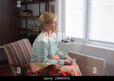 Una triste anziana donna caucasica siede su una sedia sotto una coperta vicino alla finestra con un riscaldatore vicino ai piedi e tiene i soldi nelle sue mani. Prezzi in aumento per Foto Stock