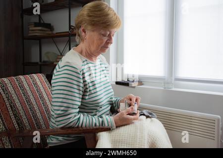 Una donna anziana siede su una sedia sotto una coperta vicino alla finestra con un riscaldatore vicino ai piedi e tiene i soldi nelle sue mani. Aumento dei prezzi dell'energia Foto Stock