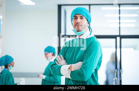 Medici professionisti e team sorridenti mentre si trova in un corridoio ospedaliero con un gruppo diversificato di personale sullo sfondo Foto Stock