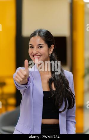 Donna d'affari radiosa in un blazer lilla che regala il pollice in un ambiente informale con un sorriso travolgente. Foto Stock