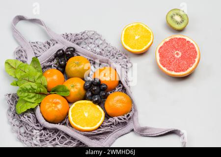 Sacchetto in rete riutilizzabile con frutta. Mandarini, pompelmo, kiwi, uva e foglie di bietola in tavola. Sfondo grigio. Posizione piatta Foto Stock