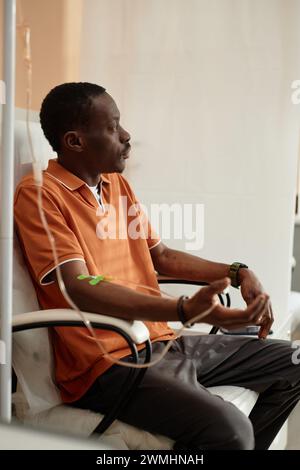 Verticale vista laterale ritratto di pensivo afroamericano uomo ottenere IV trattamento di gocciolamento seduto in sedia con tubi medici alla clinica Foto Stock