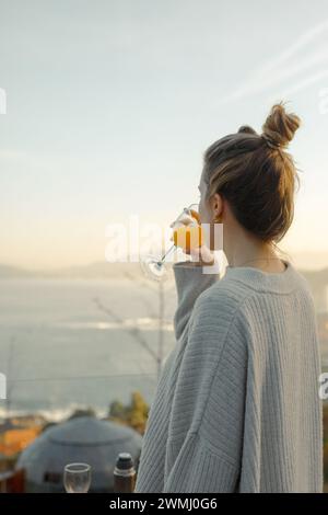 Eleganza mattutina: Giovane donna che si gusta un fresco bicchiere di succo d'arancia su una terrazza affacciata sul mare all'alba Foto Stock