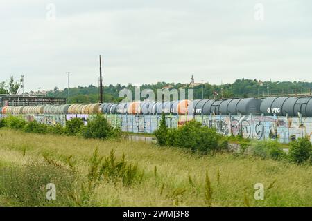 Lione, Francia - 16 maggio 2023: Un lungo treno merci si erge sui binari in una giornata nuvolosa, in primo piano c'è una recinzione ricoperta di graffiti luminosi Foto Stock