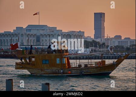 Scatto al tramonto sulla cornica di Doha che mostra il Dhow nel golfo arabo con la bandiera del Qatar che sventolava, auto per strada, persone che camminano e Amiri Diwan sullo sfondo Foto Stock