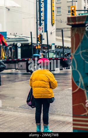 Vancouver, Canada - 21 febbraio 2024: Una donna vestita con una giacca gialla brillante e un berretto rosso brillante, in attesa di attraversare Georgia Street. Foto Stock