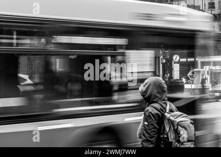 Vancouver, Canada - 21 febbraio 2024: Una persona è in attesa di attraversare Georgia Street, con un autobus che passa, il suo movimento è offuscato dalla velocità dell'otturatore. Foto Stock