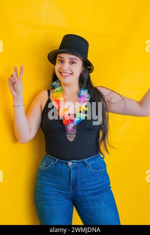 Giovane donna caucasica con un cappello nero e una collana di fiori, festeggiando con un grande sorriso. sfondo giallo Foto Stock