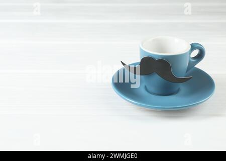 Tazza con baffi di carta finti e piattino su un tavolo di legno bianco, spazio per il testo Foto Stock