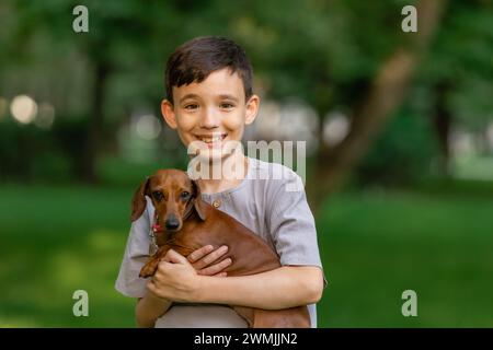 un ragazzo carino tiene un cane da dachshund tra le braccia durante una passeggiata estiva. Foto di alta qualità Foto Stock