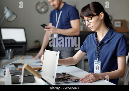 Giovane ingegnere donna del centro di assistenza riparazione che guarda al computer portatile smontato e identifica il problema tecnico in base al luogo di lavoro Foto Stock