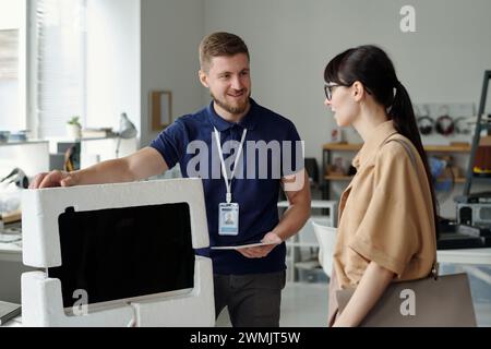 Giovane e sorridente manager del centro di risoluzione dei problemi che tiene la mano del monitor del computer imballato e lo restituisce al cliente dopo la riparazione Foto Stock
