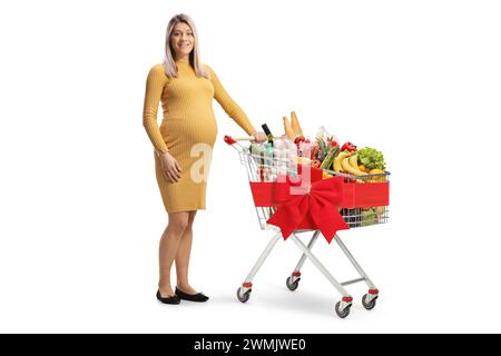 Donna incinta in piedi con un carrello pieno di generi alimentari legato con un nastro rosso isolato su sfondo bianco Foto Stock