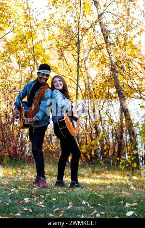 Coppia di sposi in corsa mista che si guardano a vicenda sorridendo e suonando le chitarre durante una gita in famiglia autunnale in un parco cittadino, girano... Foto Stock