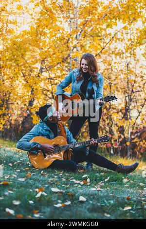 Coppia di sposi in gara mista che trascorrono del tempo insieme, sorridendo e suonando le chitarre durante una gita in famiglia autunnale in un parco cittadino Foto Stock