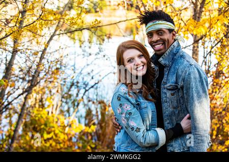 Una coppia di sposi in gara mista che sorride alla telecamera, trascorrendo del tempo insieme durante una gita in famiglia autunnale in un parco cittadino Foto Stock