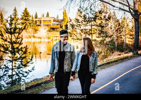 Una coppia di sposi di razza mista che si tiene per mano e sorride l'un l'altro, che cammina lungo una strada in un parco cittadino durante una gita di famiglia autunnale, spendendo quali... Foto Stock