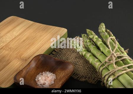 Mazzo di asparagi verdi legato con filo di iuta vicino al tagliere su sfondo nero con spazio copia. Cottura degli asparagi e concep di verdure primaverili Foto Stock