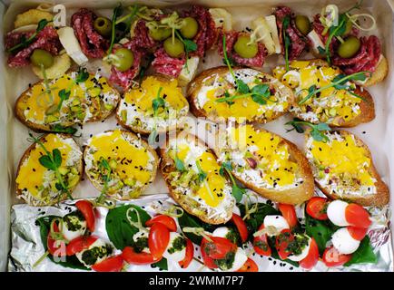 Bruschette condite con avocado e formaggio, accompagnate da tartine adornate con pomodori ciliegini, mozzarella, pesto, salame, formaggio, e olive Foto Stock