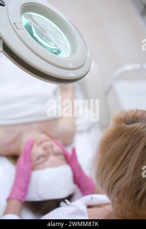 Un cliente si rilassa sotto una lampada con lente d'ingrandimento illuminata durante un esame della pelle presso una clinica di bellezza, con un cosmetologo a fuoco Foto Stock