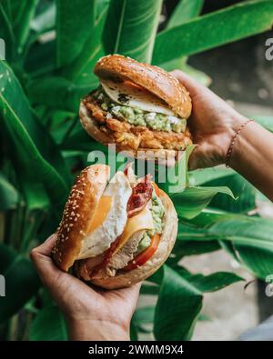 Le mani delle persone tengono in mano due bagel di hamburger pieni di uova, verdure, pollo, salsa e formaggio, dietro ci sono le piante Foto Stock