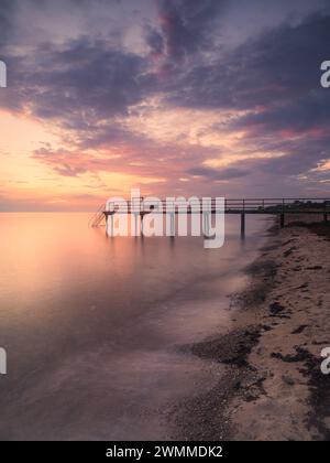 Un tramonto sereno proietta un caldo bagliore su un mare tranquillo accanto ad un molo di legno in Svezia. Il molo si estende nelle acque tranquille, sotto un cielo dipinto Foto Stock