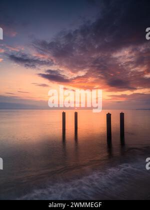 Un tranquillo tramonto proietta calde sfumature nel cielo, riflettendo sulle calme acque di una spiaggia di mare svedese. Quattro pali di legno si trovano silenziosamente nel mosto Foto Stock