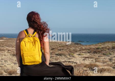 Vista posteriore di una giovane e graziosa donna con lunghi capelli rossi che indossa un abito nero e uno zaino giallo brillante sulla schiena mentre guarda le terre costiere Foto Stock