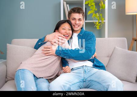 Felice giovane coppia multirazziale che si abbraccia seduto sul divano nel soggiorno. Foto Stock