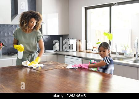 Madre e figlia birazziali puliscono la cucina insieme a casa per figlia. Condividono un momento di lavoro di squadra in un ambiente domestico luminoso. Foto Stock