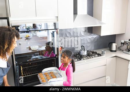 La madre birazziale si fa cuocere con sua figlia a casa per sua figlia. Condividono un momento di gioia per preparare cupcake in una cucina moderna. Foto Stock