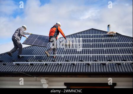 Lavoratori che trasportano pannelli solari su un tetto di casa. Due ingegneri installano una batteria solare nelle giornate di sole. Risorse energetiche alternative. Foto Stock