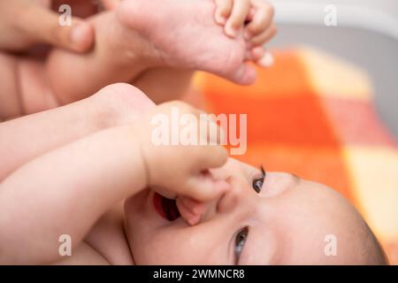 piccola bambina sorridente sdraiata su una coperta che le mette i piedi in bocca a causa del disagio causato dalla dentizione. concetto di apparizione dei primi denti Foto Stock