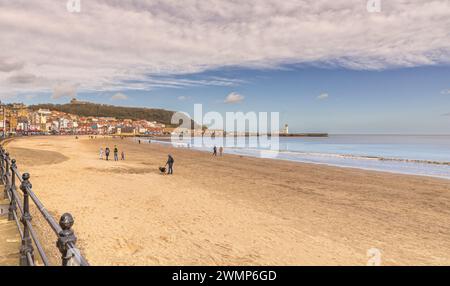 Le onde si infrangono dolcemente verso una spiaggia e un faro sorge su un molo che si estende fino al mare. Le persone sono sulla spiaggia e sulle ringhiere del pr Foto Stock