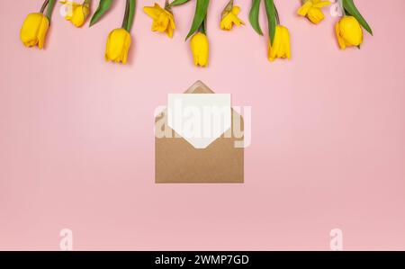 Vista dall'alto di tulipani gialli e narcisi su sfondo rosa. Busta Kraft, biglietto bianco. Composizione colorata primaverile, disposizione piatta, spazio di copia. Foto Stock