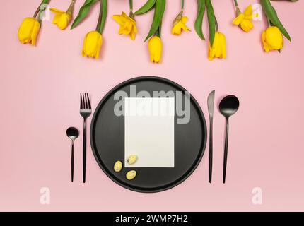 Vista dall'alto di tulipani gialli, narcisi, uova di pasqua su sfondo rosa. Tavolo, piatto nero e posate, carta bianca. Composizione primaverile di pasqua. Foto Stock